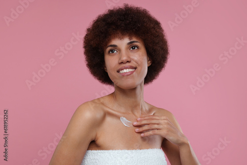 Beautiful young woman applying cream onto body on pink background