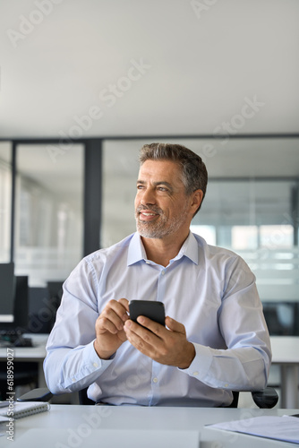 Mid aged mature happy confident business man ceo executive manager holding smartphone using mobile apps on cell phone sitting at work desk in office looking away and thinking, vertical.