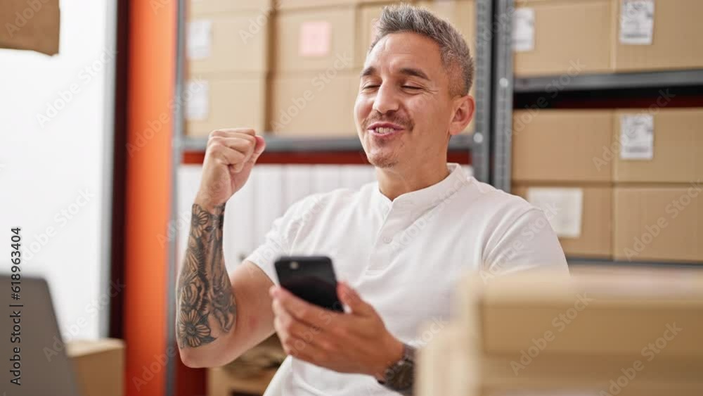 Young hispanic man ecommerce business worker using smartphone celebrating at office Young hispanic man ecommerce business worker using smartphone celebrating at office