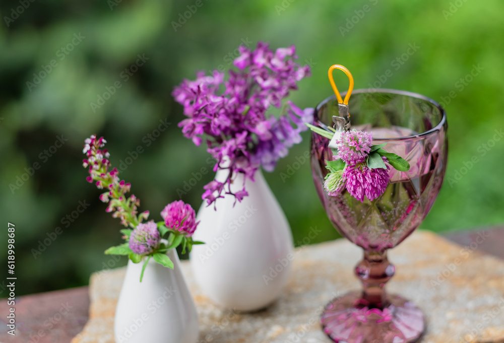 A glass with a drink and flowers in a white vase on the terrace against the backdrop of a green garden A glass with a drink and flowers in a white vase on the terrace against the backdrop of a green garden