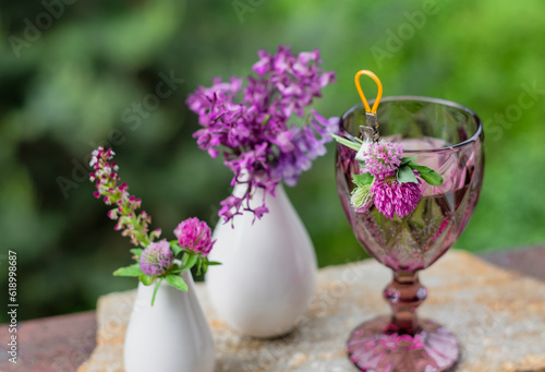 A glass with a drink and flowers in a white vase on the terrace against the backdrop of a green garden
