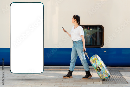 Side view of young woman walking with her luggage using cellphone. Tarin in background. Big smartphone with mock up and copy space. Concept of travel and business trip. photo