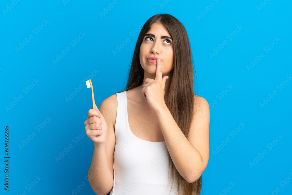 Young caucasian woman brushing teeth isolated on blue background having doubts while looking up Young caucasian woman brushing teeth isolated on blue background having doubts while looking up