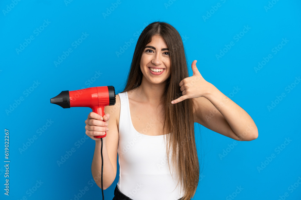 Young caucasian woman holding a hairdryer isolated on blue background making phone gesture. Call me back sign Young caucasian woman holding a hairdryer isolated on blue background making phone gesture. Call me back sign