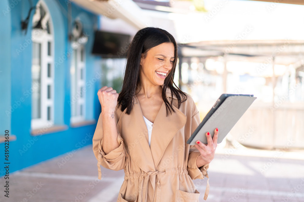 Fototapeta premium Young woman at outdoors holding a tablet and celebrating a victory