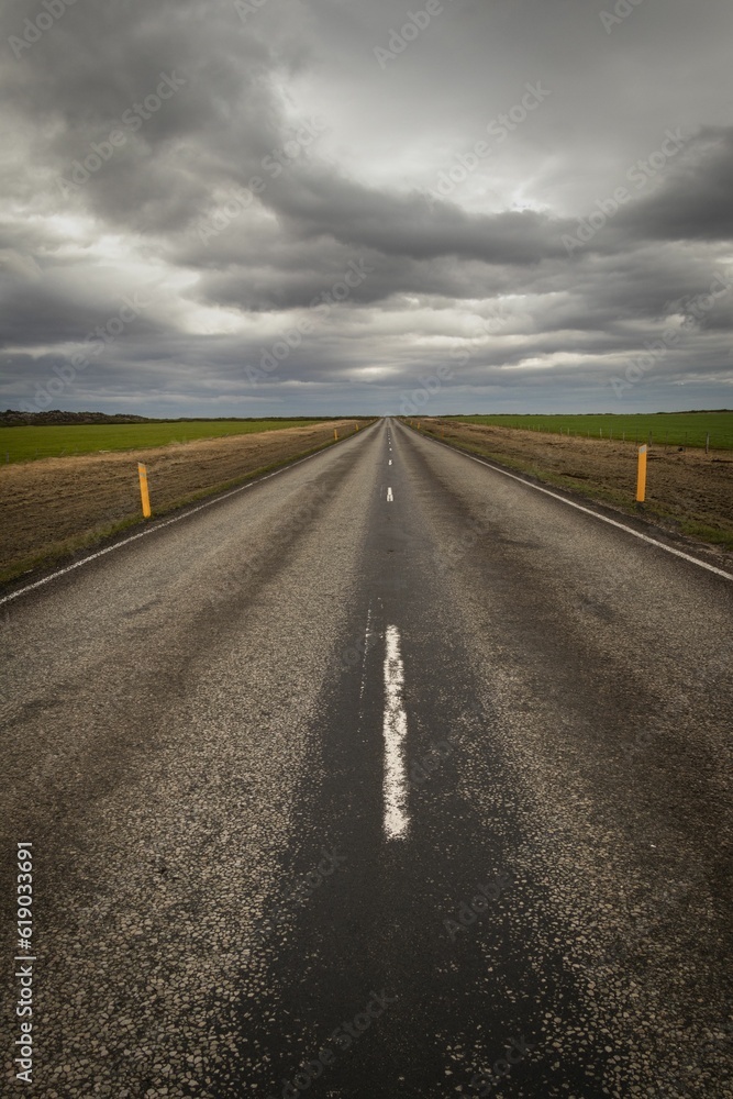 Vertical shot of a road stretching through a rolling landscape of grassy fields beneath a cloudy sky