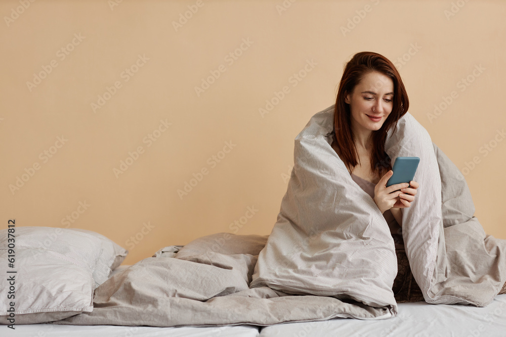 Minimal portrait of smiling young woman sitting on comfortable bed wrapped in blanket and holding smartphone scrolling social media, copy space