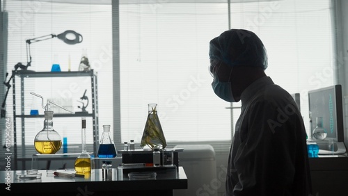 A male scientist examines an Erlenmeyer flask with a plant inside. Dark silhouette of a scientist in a white coat, cap, goggles and a mask in the laboratory. Scientific experiment.