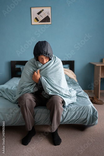 Senior man freezing indoors without central heating. Old male wearing warm winter clothes sitting on bed in very cold room at home with tired disappointed face expression