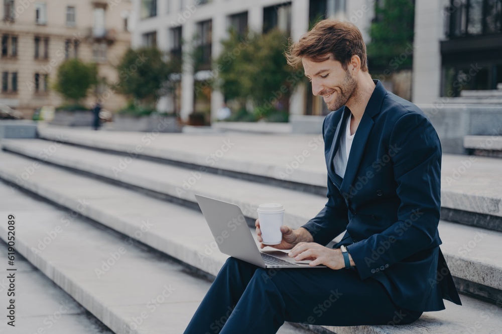 Satisfied businessman in blue suit works remotely on laptop outside office, enjoying sunny lunch break. Satisfied businessman in blue suit works remotely on laptop outside office, enjoying sunny lunch break.