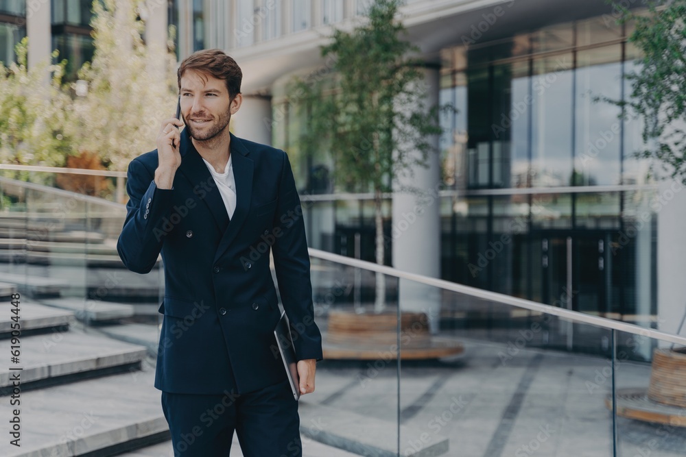 Fashionable businessman uses tech, has phone call, holds gadget, poses near office. Economist makes focused phone call. Fashionable businessman uses tech, has phone call, holds gadget, poses near office. Economist makes focused phone call.