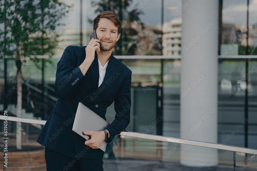 Confident bearded businessman in formal attire makes business call outside glass building, laptop in relaxed pose, talking to colleagues. Confident bearded businessman in formal attire makes business call outside glass building, laptop in relaxed pose, talking to colleagues.