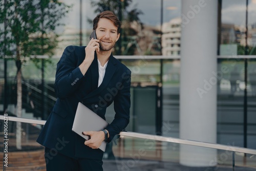 Confident bearded businessman in formal attire makes business call outside glass building, laptop in relaxed pose, talking to colleagues.
