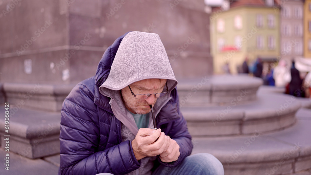 Adult man в очках sitting on square and lights up at a tobacco pipe in the Palace Square, Warsaw Old Town Adult man в очках sitting on square and lights up at a tobacco pipe in the Palace Square, Warsaw Old Town