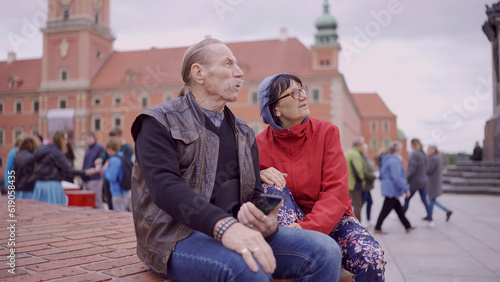 Elderly couple of tourists are sitting and talking in the historic center of an old European city  senior has a smartphone in his hand. Palace Square  Warsaw Old Town