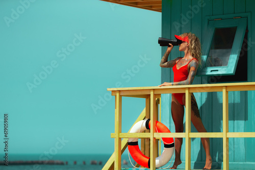 Lifeguard woman in red bikini and visor with binocular and lifebuoy on watch tower against turquoise sky