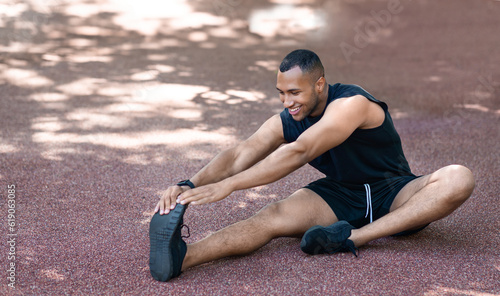 Happy handsome african american guy in sportswear stretching leg muscles for jogging at park