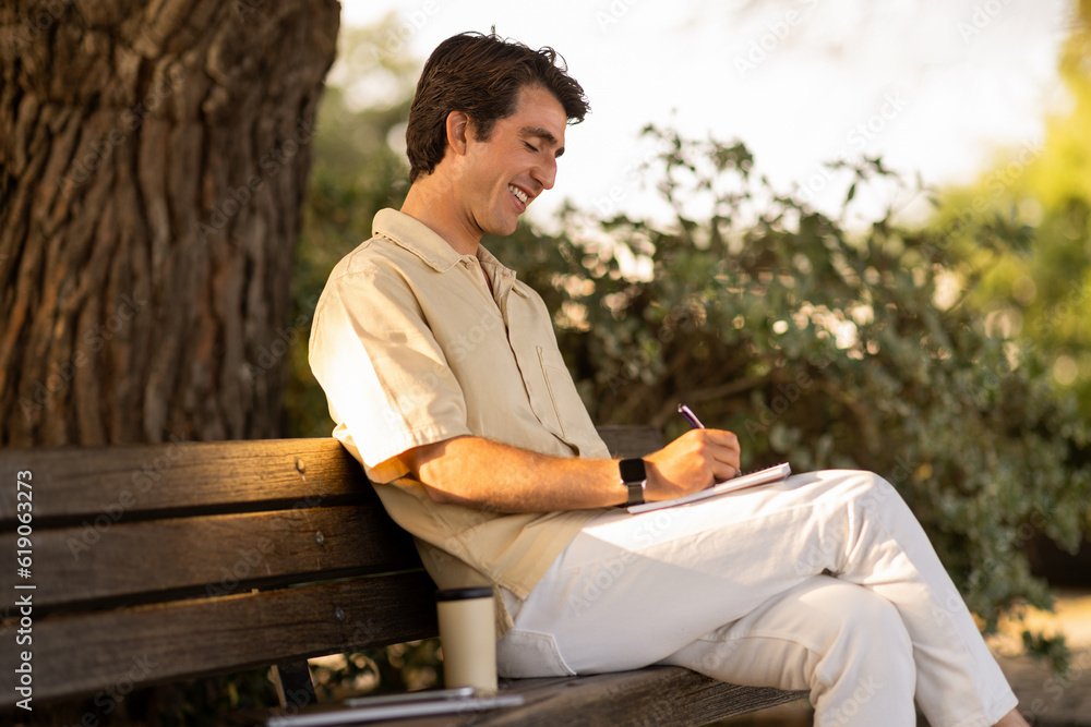 Creative man writer sitting on bench at park, taking notes