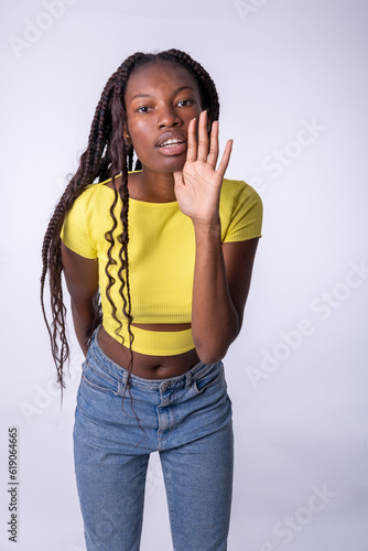 Mujer africana parada sobre fondo blanco con la mano en la boca contando rumores secretos, susurrando una conversación maliciosa