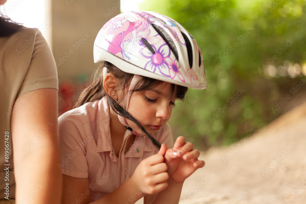 Little dark-haired girl cyclist in a helmet sits sadly outdoors Little dark-haired girl cyclist in a helmet sits sadly outdoors
