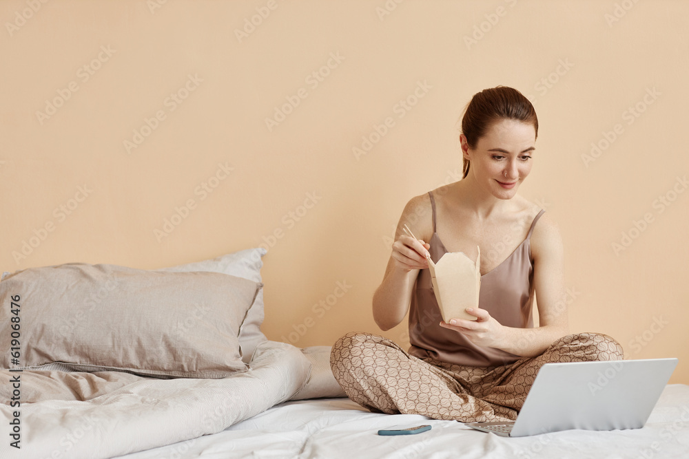 Minimal portrait of smiling young woman eating takeout noodles and watching videos via laptop enjoying relaxing weekend at home, copy space Minimal portrait of smiling young woman eating takeout noodles and watching videos via laptop enjoying relaxing weekend at home, copy space