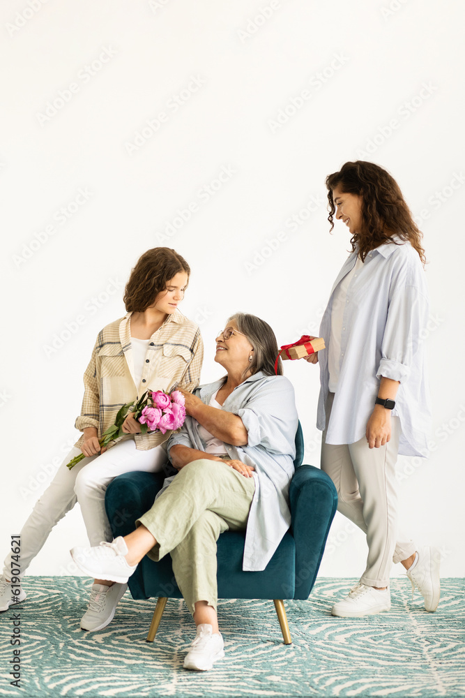 Cheerful european adult and teenager women give bouquet of flowers, gift to old grandmother