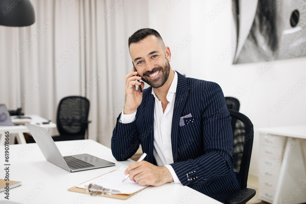 Smiling european adult guy in suit with beard works on laptop, calls by phone Smiling european adult guy in suit with beard works on laptop, calls by phone