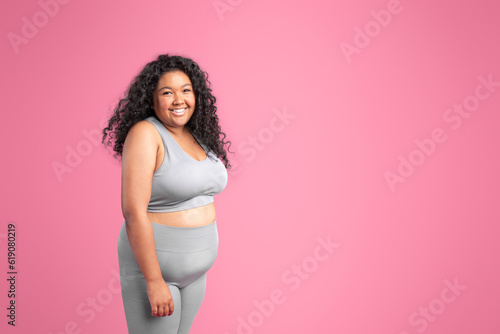 Portrait of body positive black lady in sports top bra and leggings posing at camera on pink background, free space