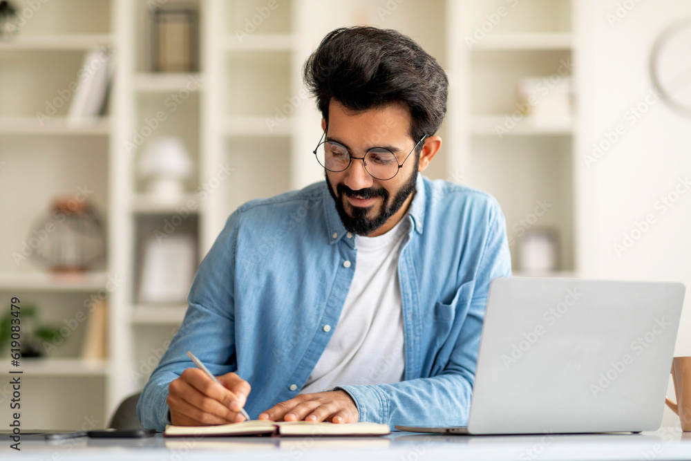 Smiling Indian Man Taking Notes While Working With Laptop At Home Office Smiling Indian Man Taking Notes While Working With Laptop At Home Office