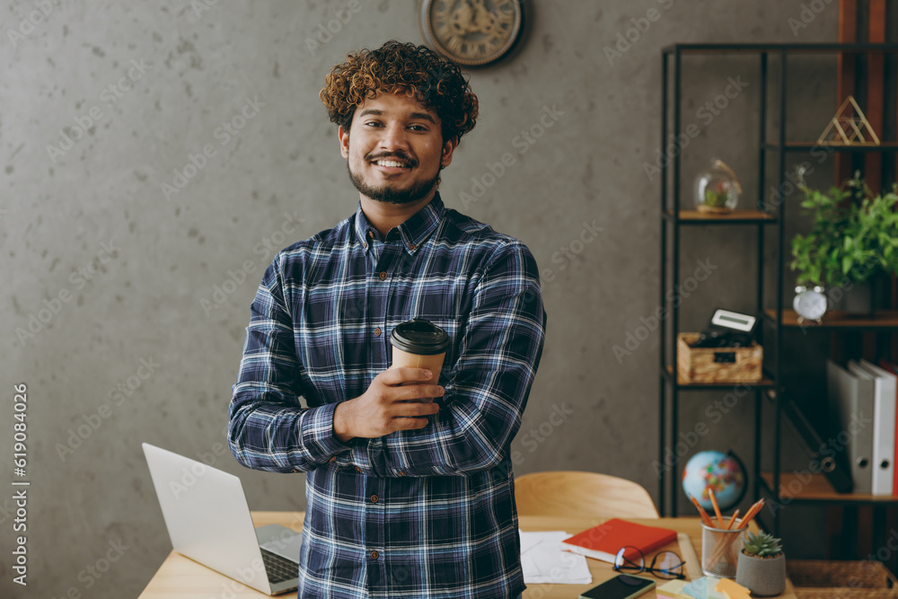 © ViDi Studio - Cheerful young employee business Indian man he wears casual blue checkered shirt hold takeaway delivery craft paper brown cup coffee to go work at office sit on desk with laptop pc computer indoors. © ViDi Studio - Cheerful young employee business Indian man he wears casual blue checkered shirt hold takeaway delivery craft paper brown cup coffee to go work at office sit on desk with laptop pc computer indoors.