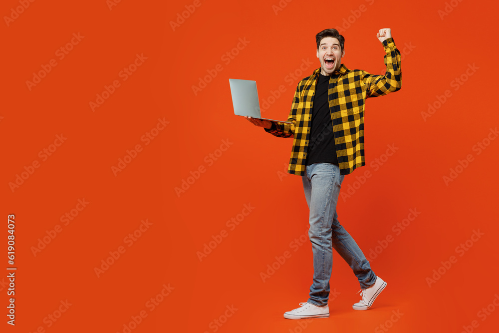 Full body side view young caucasian IT man wear yellow checkered shirt black t-shirt hold use work on laptop pc computer do winner gesture isolated on plain red orange background. Lifestyle concept. Full body side view young caucasian IT man wear yellow checkered shirt black t-shirt hold use work on laptop pc computer do winner gesture isolated on plain red orange background. Lifestyle concept.