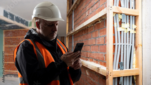 Man builder. Brigadier with phone. Architect in building under construction. Guy in uniform of builder. Engineer in helmet calls customer. Construction company contractor. Man in unfinished room