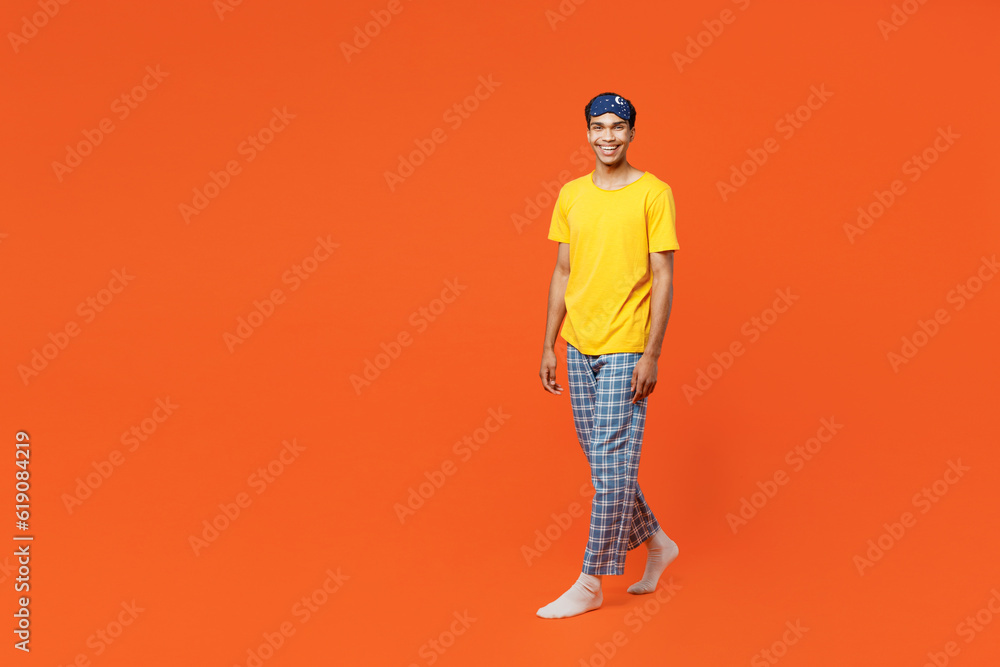 Full body smiling happy fun young man wear pyjamas jam sleep eye mask rest relax at home walking going looking camera isolated on plain orange background studio portrait. Good mood night nap concept. Full body smiling happy fun young man wear pyjamas jam sleep eye mask rest relax at home walking going looking camera isolated on plain orange background studio portrait. Good mood night nap concept.