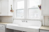 A farmhouse sink detail with white cabinets, tiled backsplash, and a chrome faucet in front of bright windows.