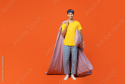 Full body smiling happy fun cheerful young man wear pyjamas jam sleep eye mask rest relax at home hold blanket pillow isolated on plain orange background studio portrait. Good mood night nap concept.