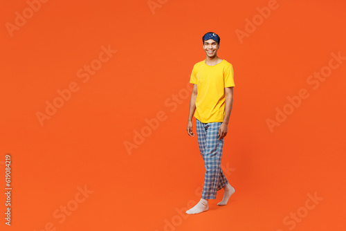 Full body smiling happy fun young man wear pyjamas jam sleep eye mask rest relax at home walking going looking camera isolated on plain orange background studio portrait. Good mood night nap concept.