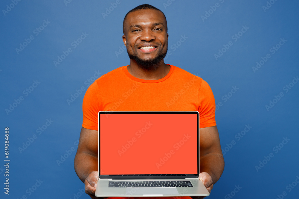 Young IT man of African American ethnicity wear orange t-shirt hold use work on laptop pc computer with blank screen workspace area isolated on plain dark royal navy blue background Lifestyle concept Young IT man of African American ethnicity wear orange t-shirt hold use work on laptop pc computer with blank screen workspace area isolated on plain dark royal navy blue background Lifestyle concept