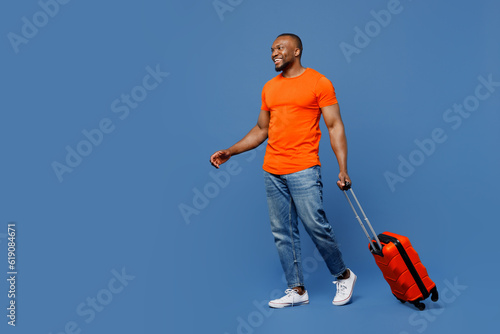 Full body traveler fun man wear orange t-shirt hold suitcase walk isolated on plain dark blue background studio. Tourist travel abroad in free spare time rest getaway. Air flight trip journey concept. #619084671
