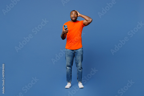 Full body fearful young man of African American ethnicity he wear orange t-shirt hold head use mobile cell phone isolated on plain dark royal navy blue background studio portrait. Lifestyle concept.