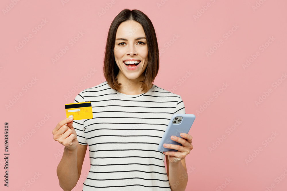 Young caucasian woman she wears casual clothes t-shirt using mobile cell phone hold credit bank card doing online shopping order delivery booking tour isolated on plain pastel light pink background. Young caucasian woman she wears casual clothes t-shirt using mobile cell phone hold credit bank card doing online shopping order delivery booking tour isolated on plain pastel light pink background.