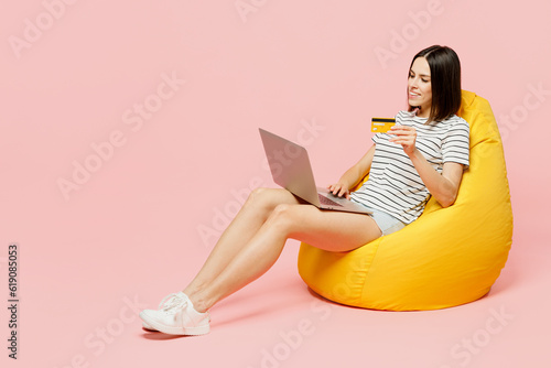 Full body young woman wears casual clothes t-shirt sit in bag chair using laptop pc computer hold credit bank card doing online shopping order delivery booking tour isolated on plain pink background.