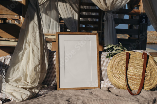 Fototapeta Naklejka Na Ścianę i Meble -  Blank picture frame, poster mockup in beach lounger, gazebo with canopy in resort at sea. Wicker straw bag with green olive tree branches in sunlight. Summer vacation, traveling concept.