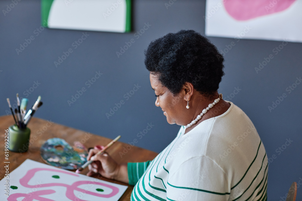 Naklejka premium Mature female artist in casualwear painting with gouache or water colors on sheet of paper while sitting by desk in studio of arts and design