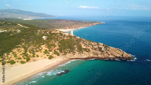 Faro de Camarinal, aerial view of the lighthouse on a cliff at the wonderful sandy beach with the blue turquoise Atlantic Ocean, Playa Los Alemanes, Andalusia, Spain