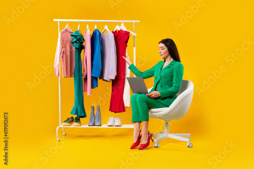 Fashion trends. Beautiful woman stylist sitting near lot of clothes on garment rack and using laptop, yellow background