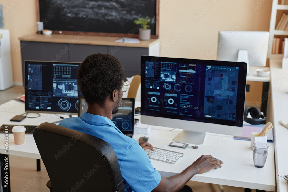 Rear view of young African American businessman sitting by workplace in front of desktop computers with graphic data on screens in office