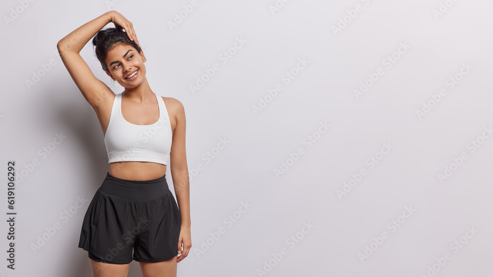 Studio shot of sporty brunette Iranian keeps arm raised up leans head stretches before workout dressed in cropped top and black shorts stands against white background copy space for your promotion Studio shot of sporty brunette Iranian keeps arm raised up leans head stretches before workout dressed in cropped top and black shorts stands against white background copy space for your promotion