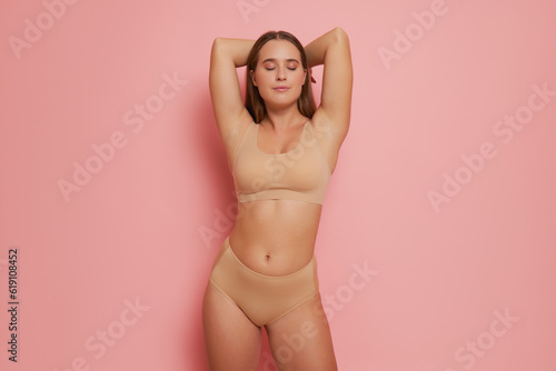 Portrait of slim girl on pink studio background, eyes closed, hands on her head, wearing beige sport bra and panties, natural beauty concept, copy space