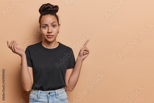 Doubtful young Latin woman raises her palm displaying clueless look points index finger on empty space showcasing your message or product dressed in casual clothes isolated over brown background