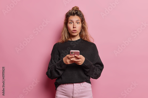Young woman with long hair reacts to shocking fake news on her phone mouth wide open in disbelief has scared expression wears casual black jumper and trousers isolated over pink studio background